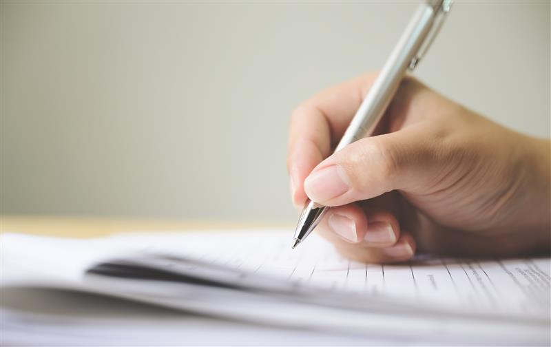 Close-up of a hand holding a silver pen, writing on a sheet of paper resting on a flat surface, with the background softly blurred.