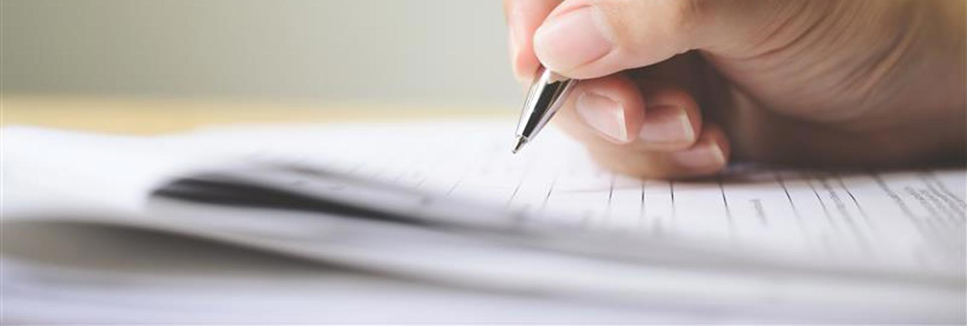 Close-up of a hand holding a silver pen, writing on a sheet of paper resting on a flat surface, with the background softly blurred.