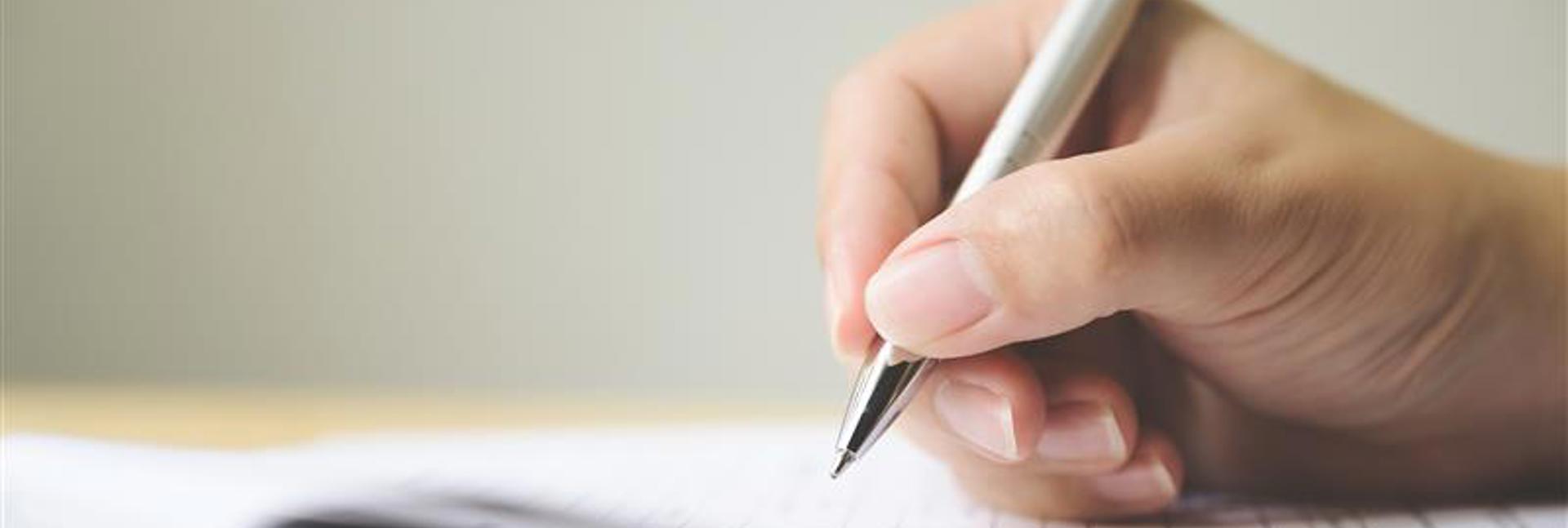 Close-up of a hand holding a silver pen, writing on a sheet of paper resting on a flat surface, with the background softly blurred.