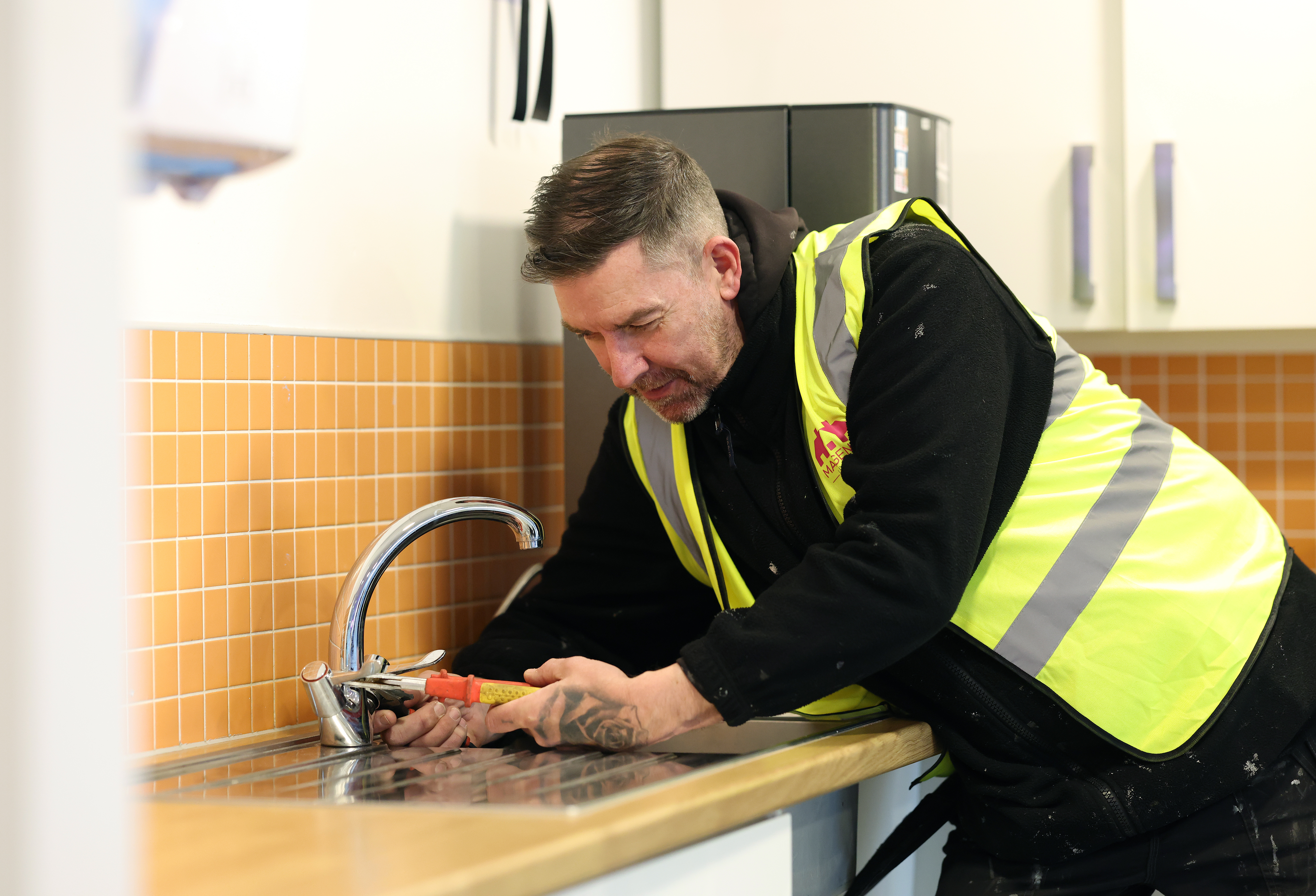 A maintenance worker wearing a high‑visibility vest uses a hand tool to fix a kitchen tap at a sink. 