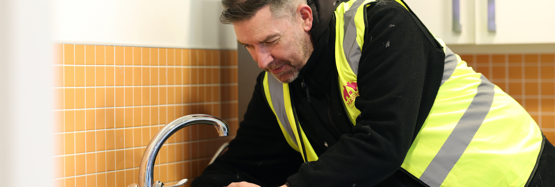 A maintenance worker wearing a high‑visibility vest uses a hand tool to fix a kitchen tap at a sink. 