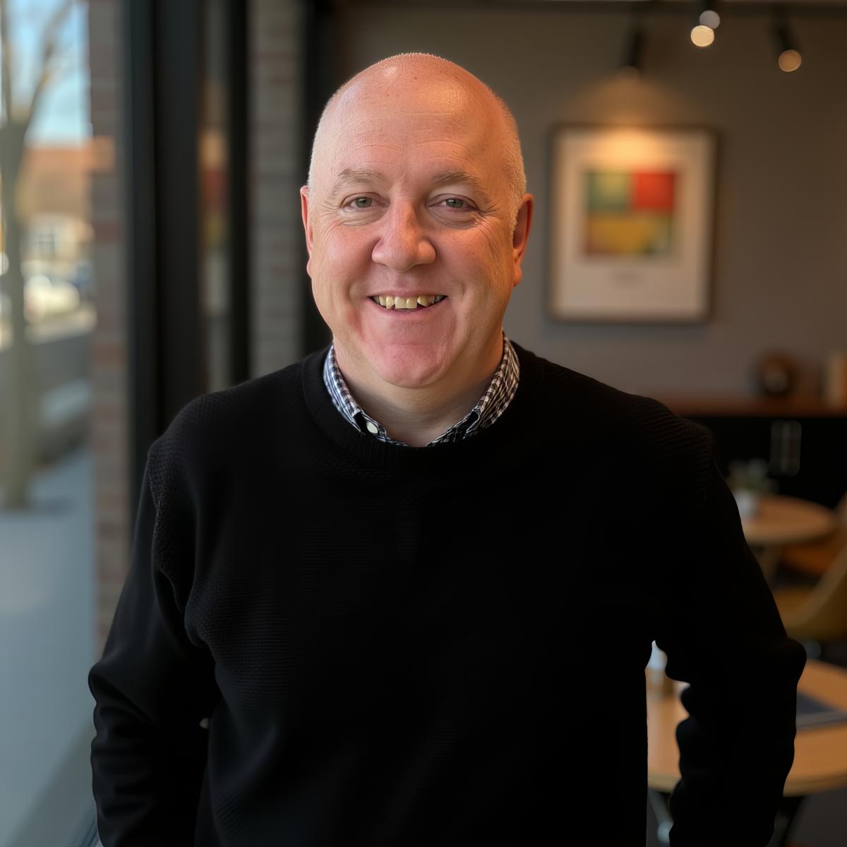 Andy Lomas standing indoors near a large window, wearing a dark jumper in a modern, softly lit room.