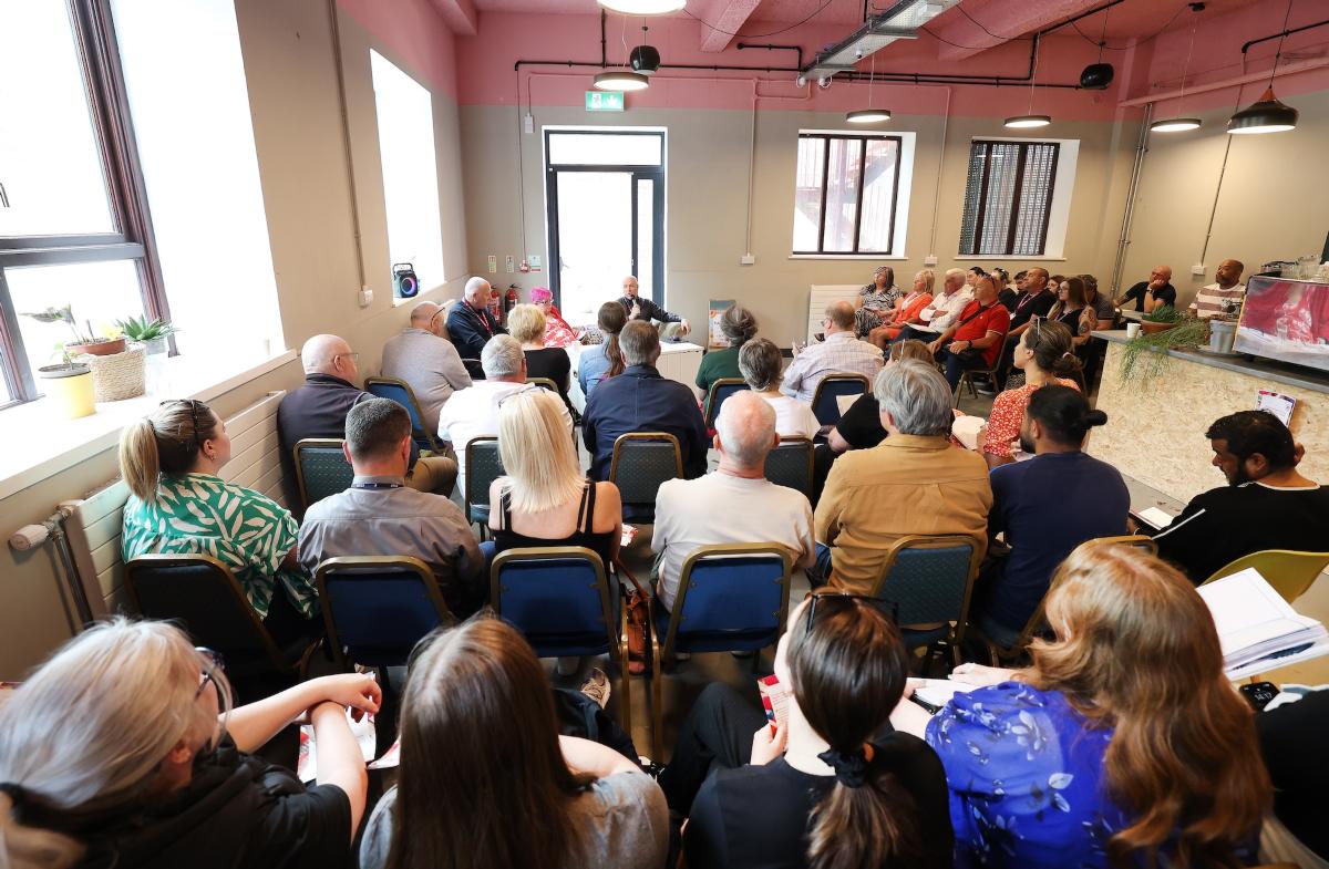 A large group of people seated in a community meeting, listening to speakers at the front of the room.