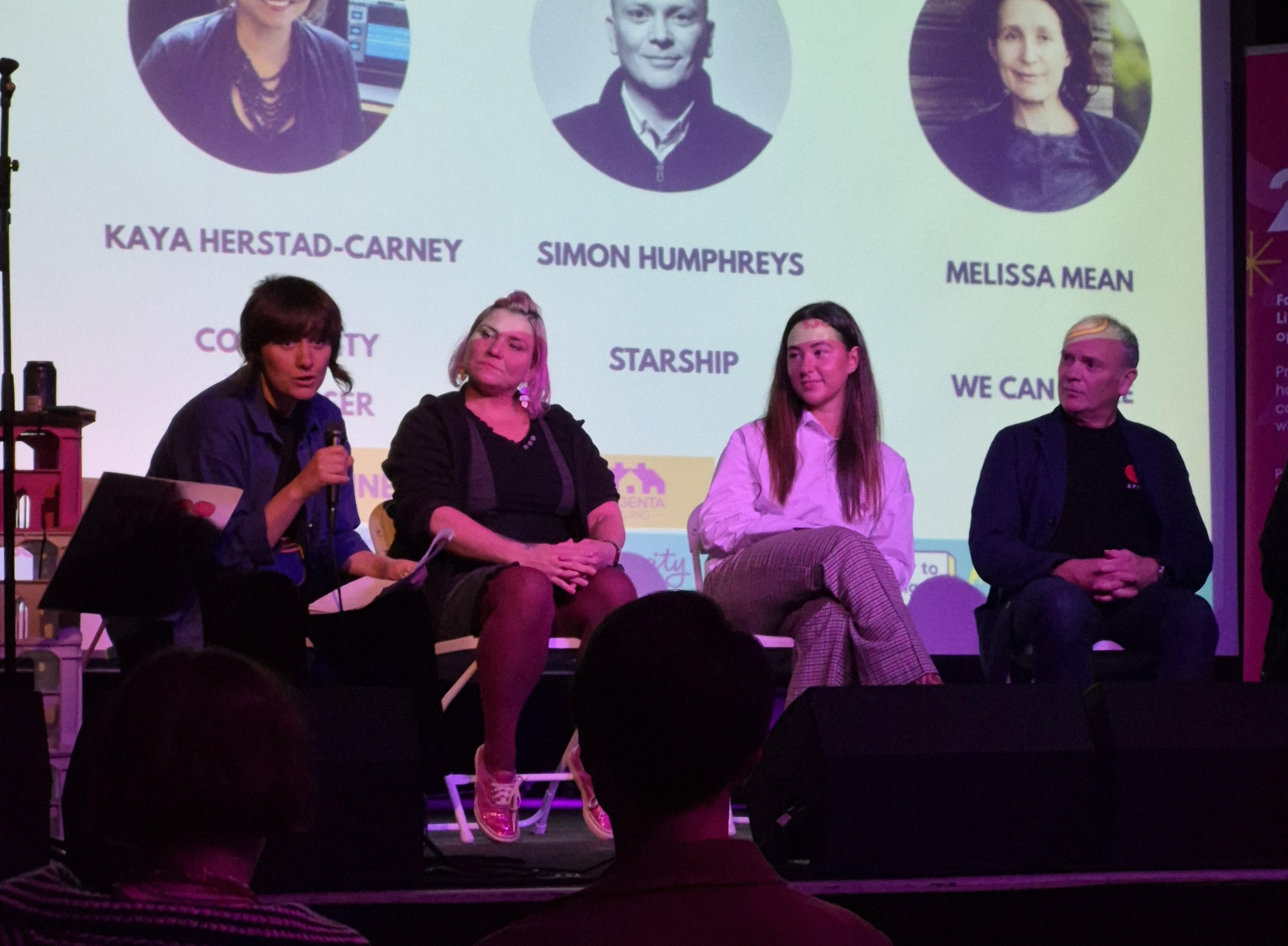 Four people seated on a stage panel, one speaking into a microphone, with a projected screen behind showing speaker names and organisations.
