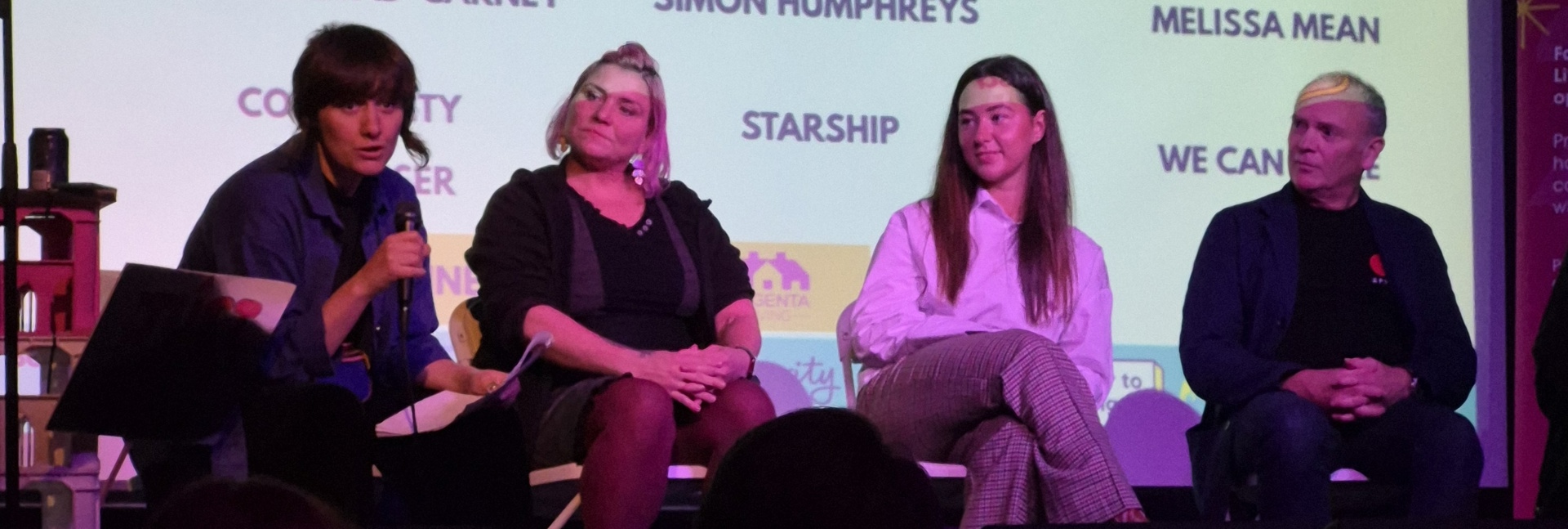 Four people seated on a stage panel, one speaking into a microphone, with a projected screen behind showing speaker names and organisations.