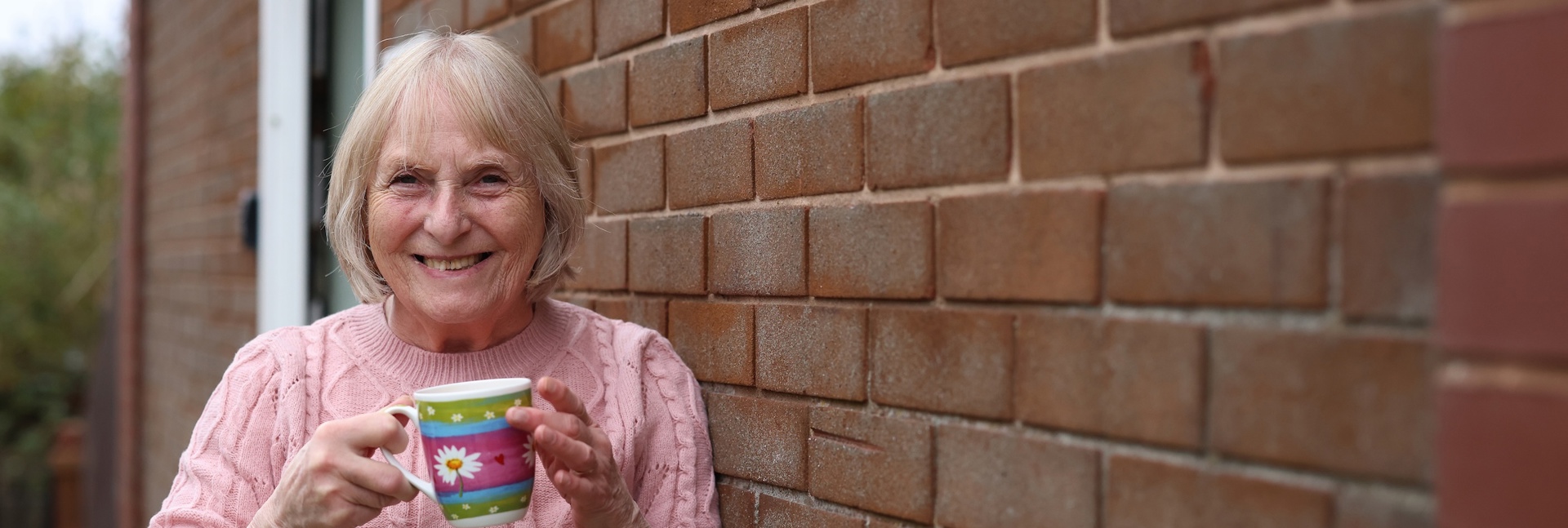 A person wearing a pink knitted jumper stands beside a brick wall outside a home, holding a colourful mug. A doorway and garden area are visible in the background, with the brick wall extending along the right-hand side of the image.