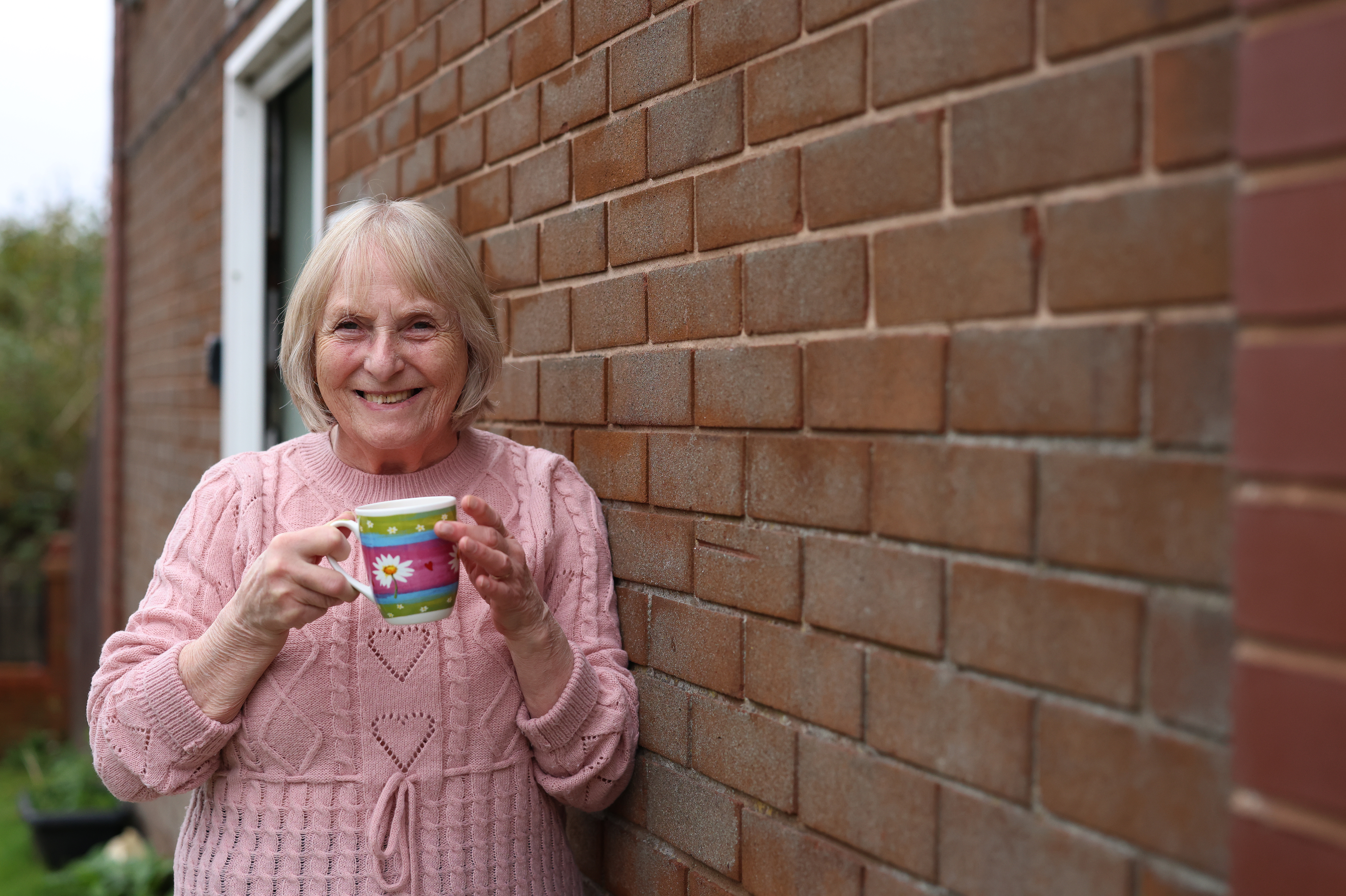 A person wearing a pink knitted jumper stands beside a brick wall outside a home, holding a colourful mug. A doorway and garden area are visible in the background, with the brick wall extending along the right-hand side of the image.