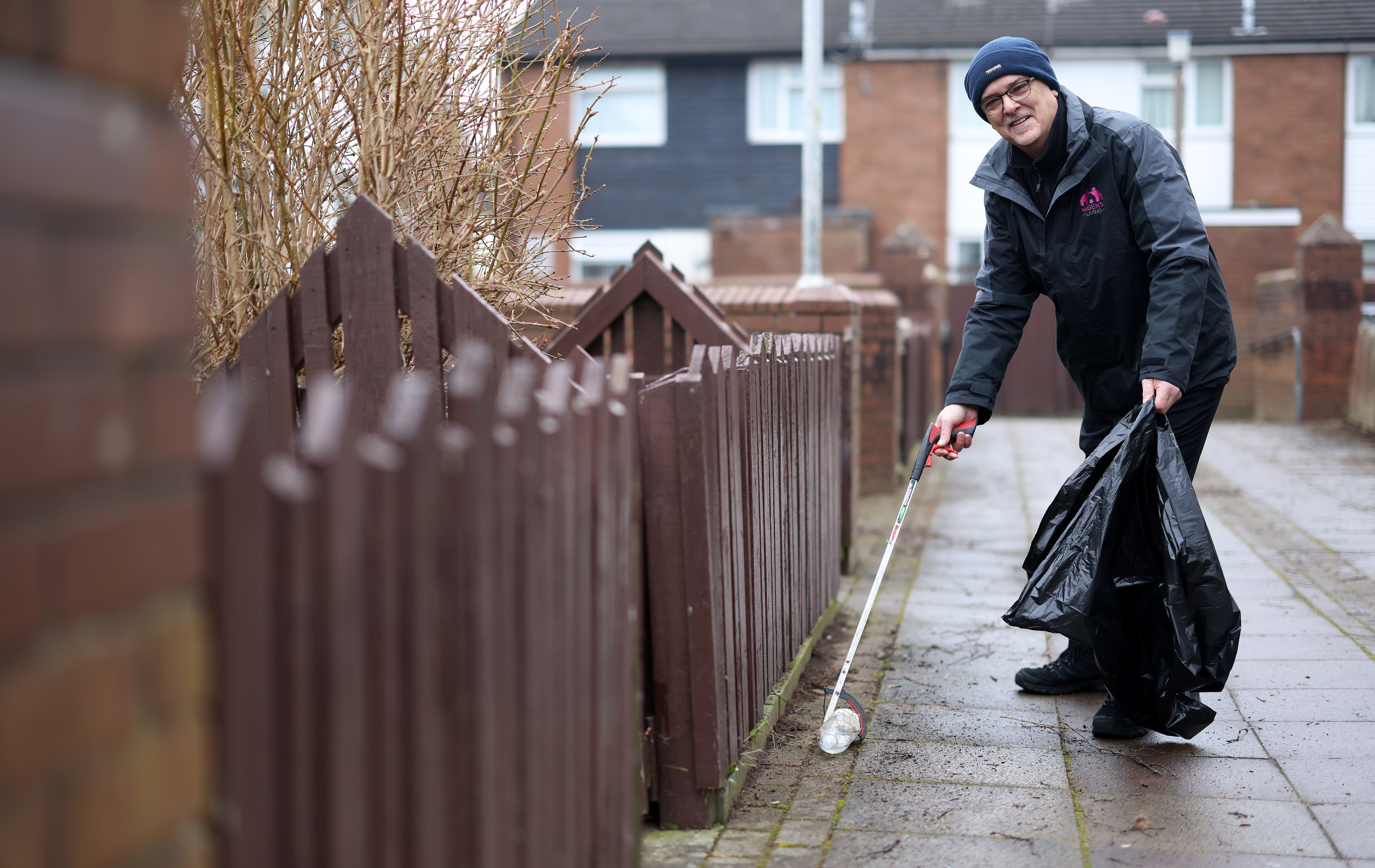 A person using a litter picker to collect rubbish along a residential pathway, holding a black bin bag.