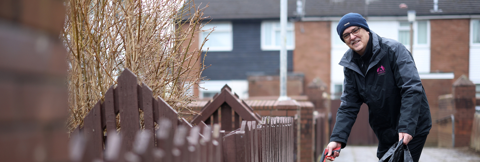 A person using a litter picker to collect rubbish along a residential pathway, holding a black bin bag.