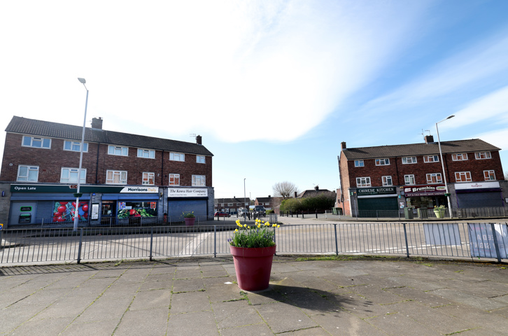 View of a neighbourhood square with shops, homes and a planter filled with flowers.