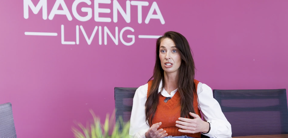 A person taking part in a conversation at a meeting table, with the Magenta Living logo visible on the wall behind them.