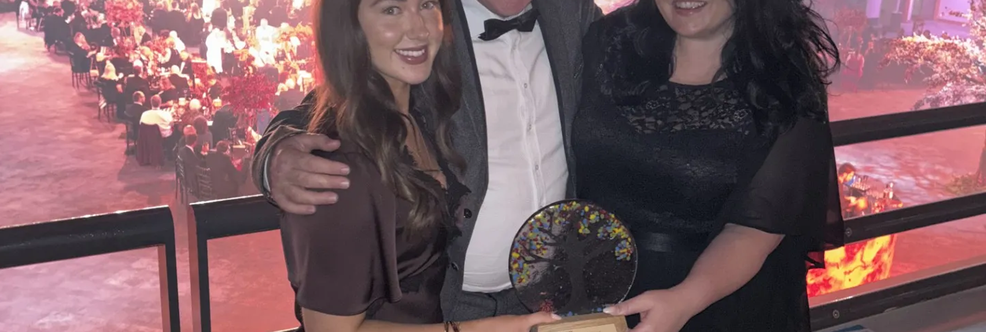 Three people in formalwear hold an award while standing on a balcony overlooking a busy, brightly lit event.