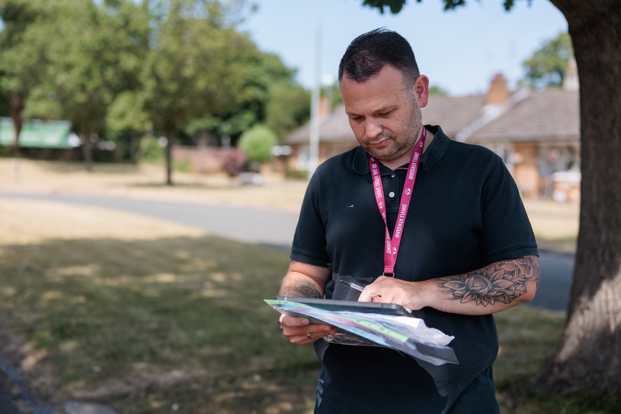 A person wearing a black polo shirt and a Magenta Living lanyard stands outdoors beneath a tree, reviewing documents on a clipboard and writing notes with a pen. The background shows a residential area with trees, grass and blurred houses in daylight.