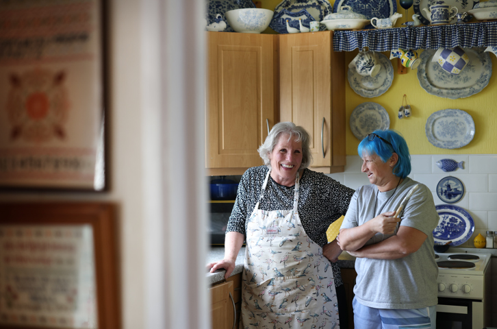 Two people talking together in a cosy kitchen with decorative plates on the wall.