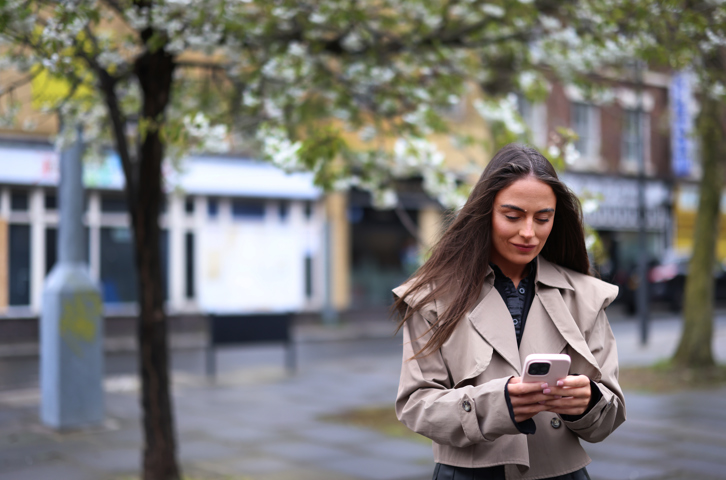 Someone using a mobile phone on a pavement with shops and trees in the background.