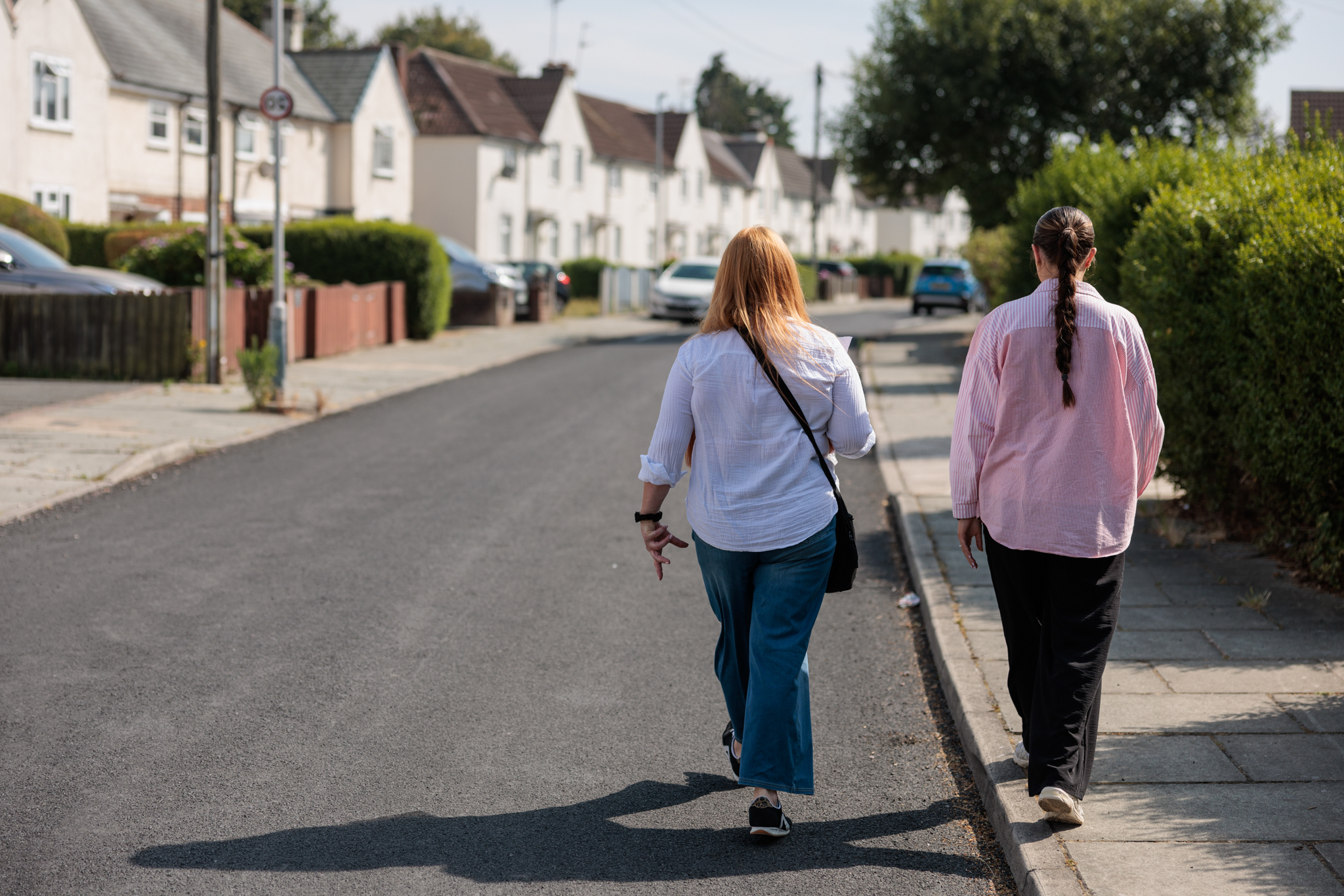Two people walking through a residential neighbourhood with houses, hedges and parked cars along the street.