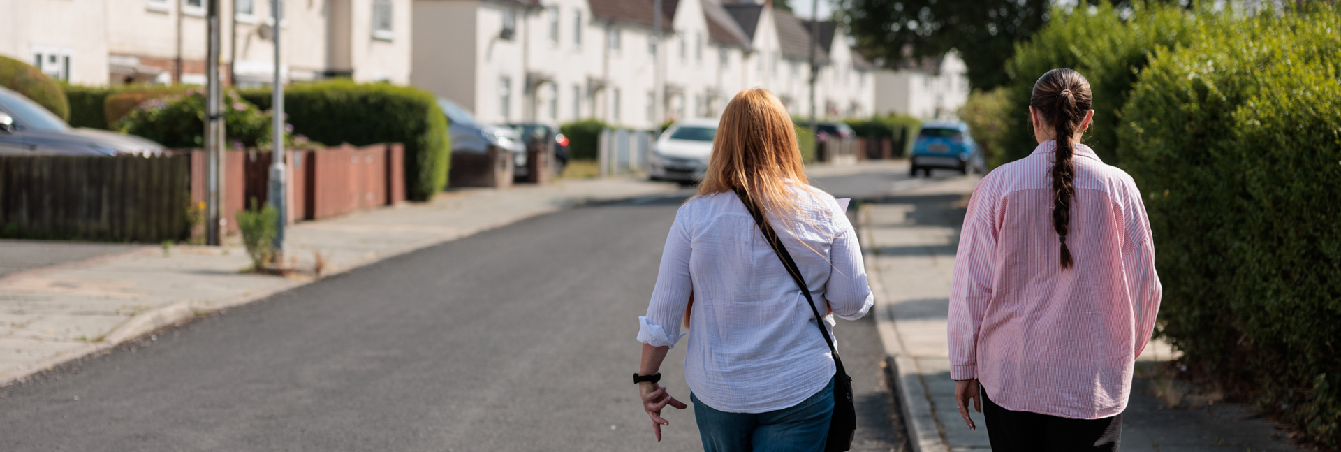 Two people walking through a residential neighbourhood with houses, hedges and parked cars along the street.