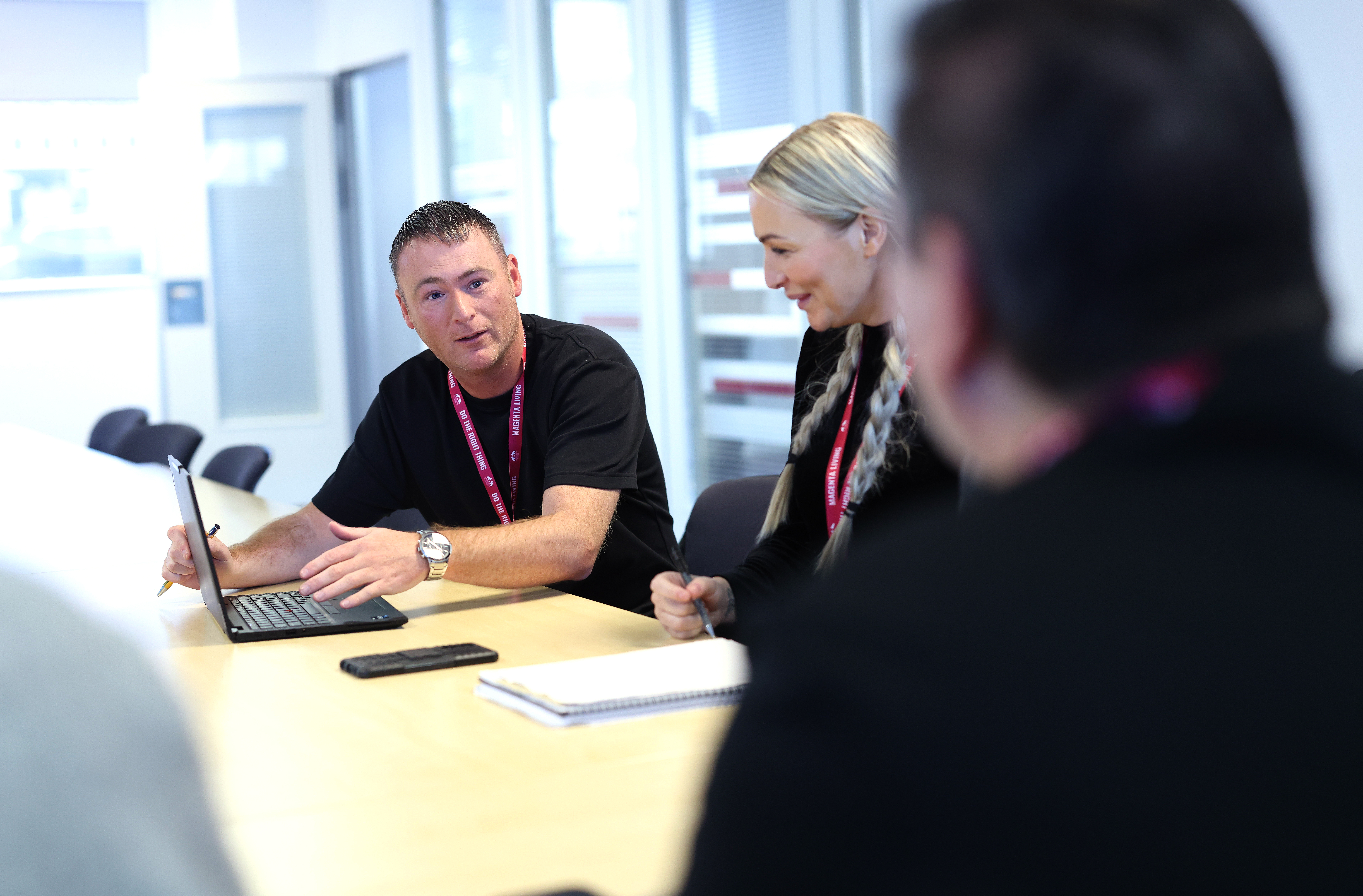 Colleagues sitting around a meeting table discussing work with a laptop and notepad.