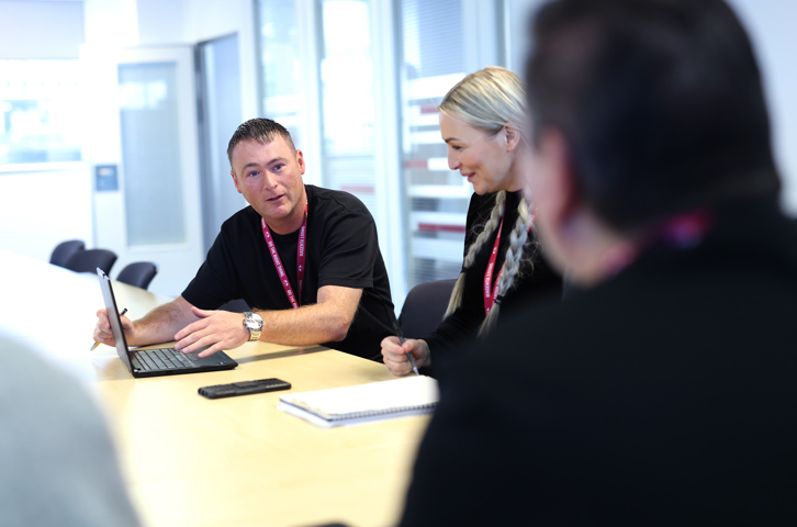 Colleagues sitting around a meeting table discussing work with a laptop and notepad.