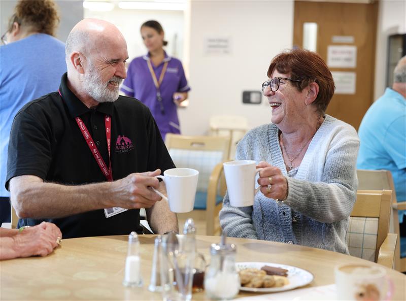 Two people sitting at a table and raising mugs together during a community coffee morning.