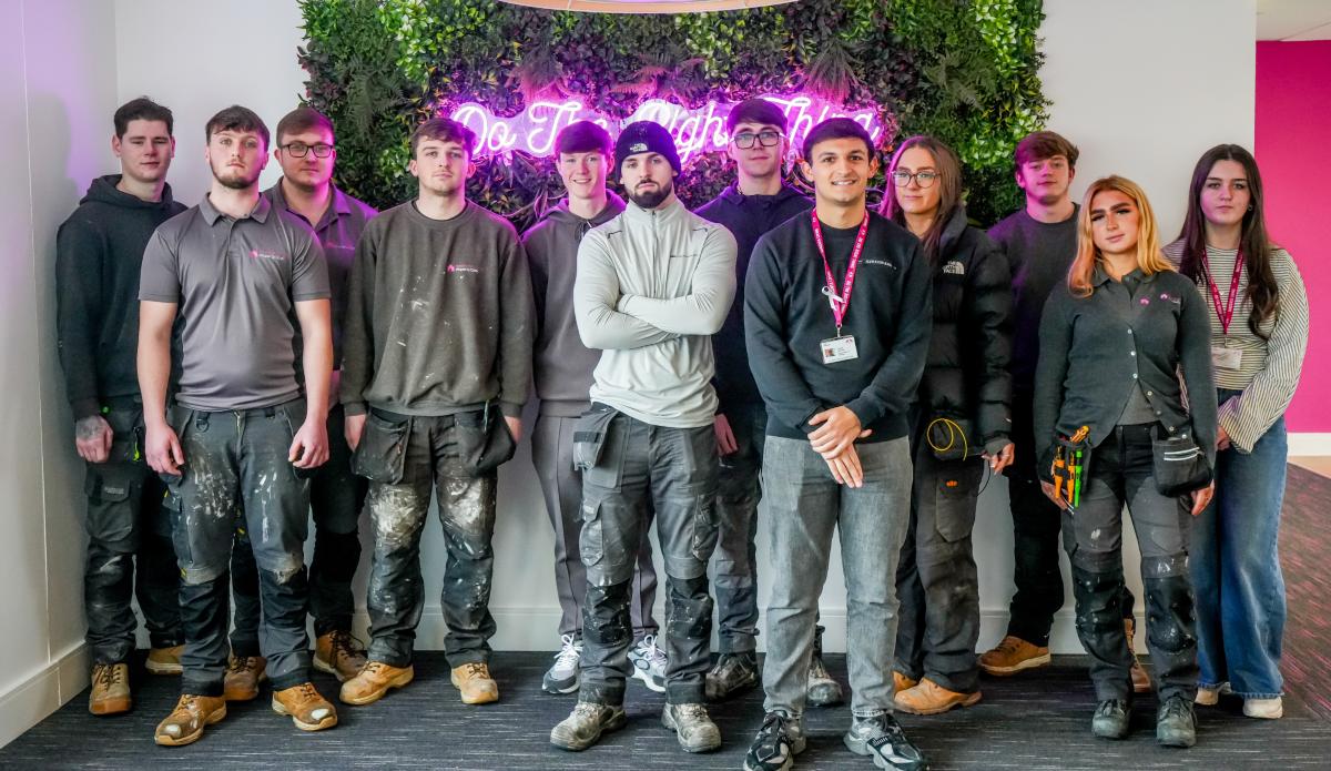 A group of apprentices standing together in front of a neon sign on a wall decorated with greenery.