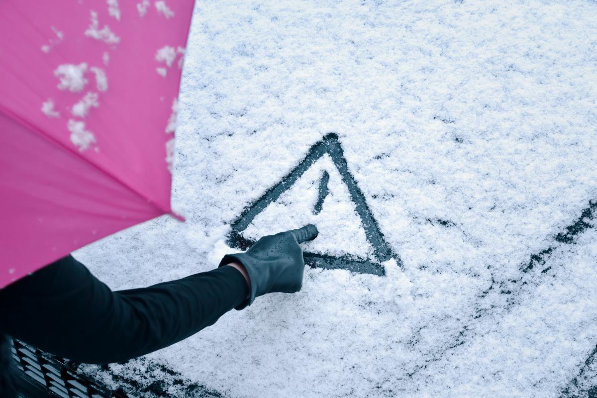 A gloved hand drawing a warning triangle in fresh snow on a car windscreen.