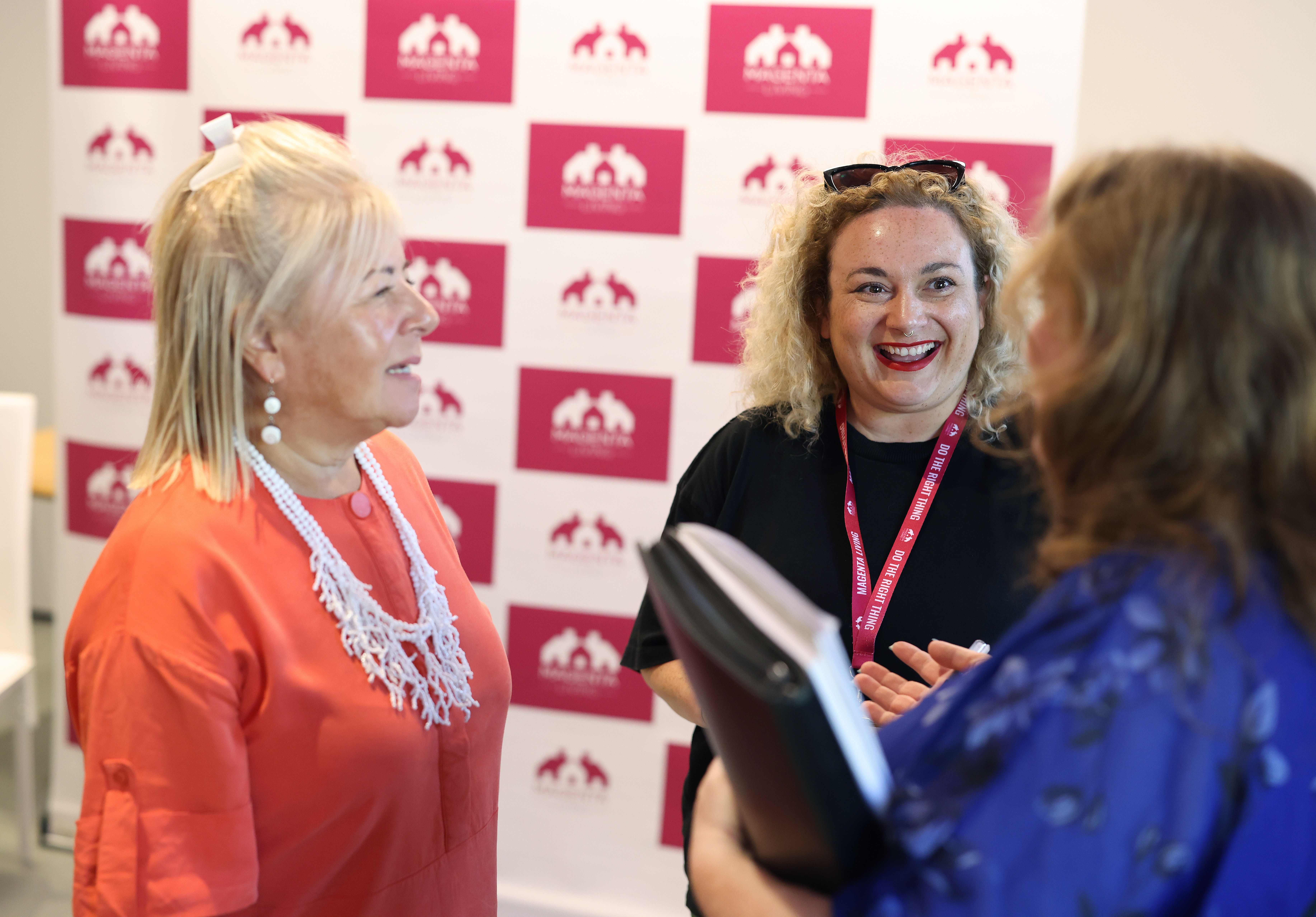 Three people talking together at an event in front of a Magenta Living backdrop.