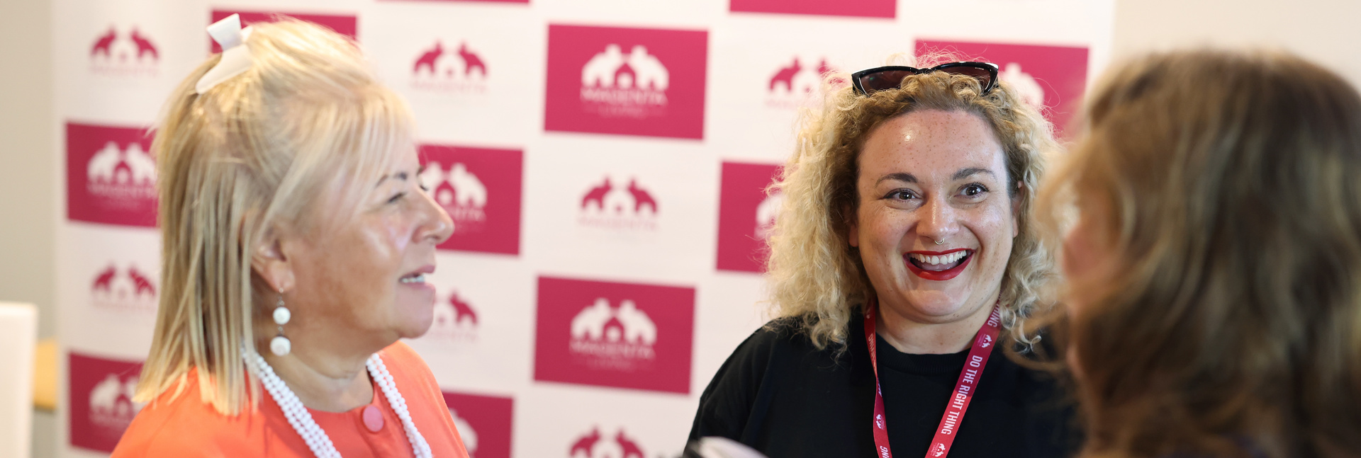 Three people talking together at an event in front of a Magenta Living backdrop.