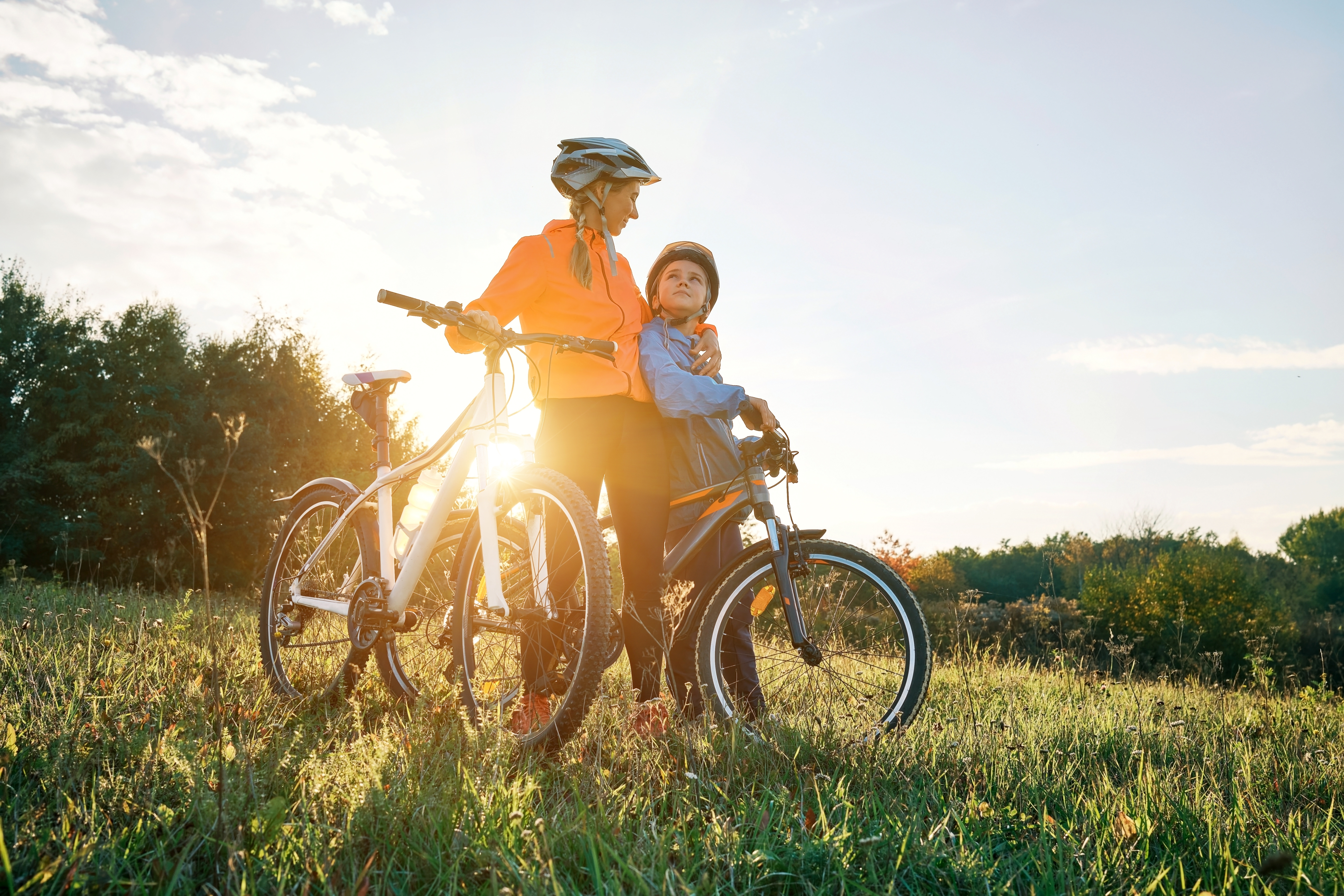 Two cyclists with helmets stand in a sunlit field holding their bikes, with trees and sky in the background.