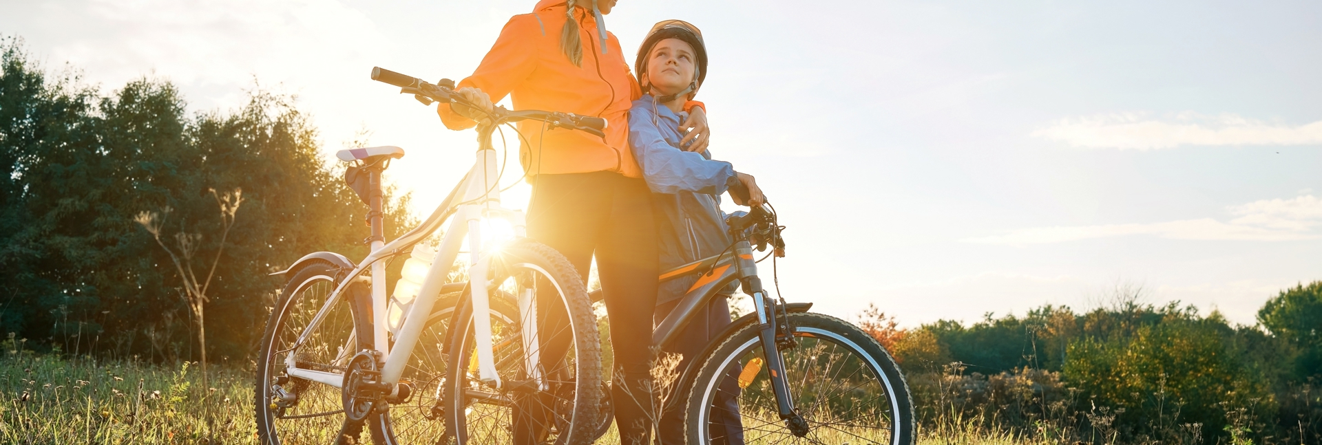 Two cyclists with helmets stand in a sunlit field holding their bikes, with trees and sky in the background.