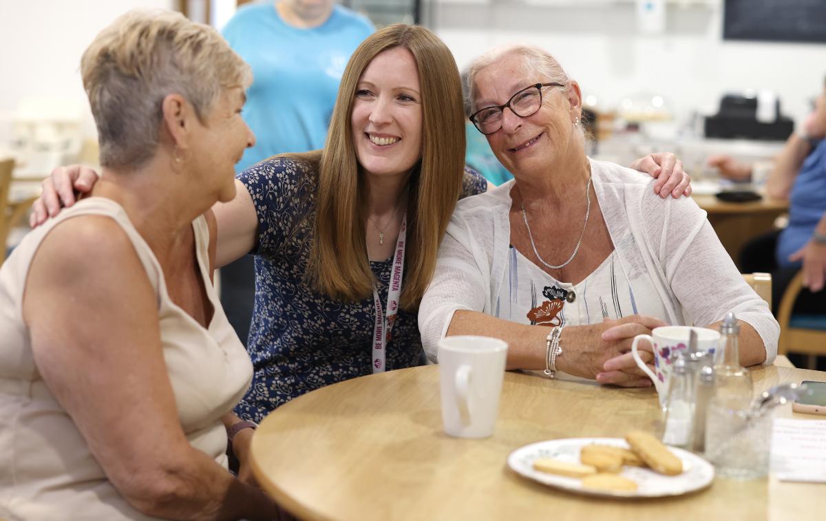 Three people sitting closely together at a community coffee morning, chatting with drinks and biscuits on the table.