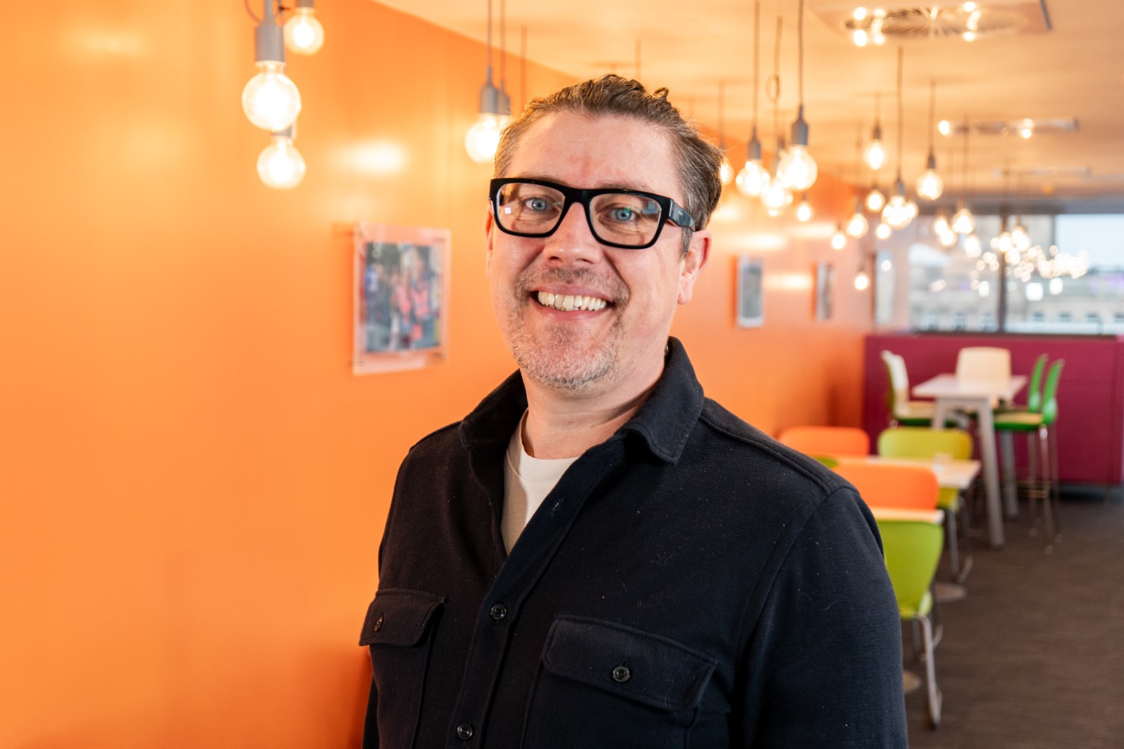 Steve Nichol standing in a bright communal area with orange walls and hanging lights.
