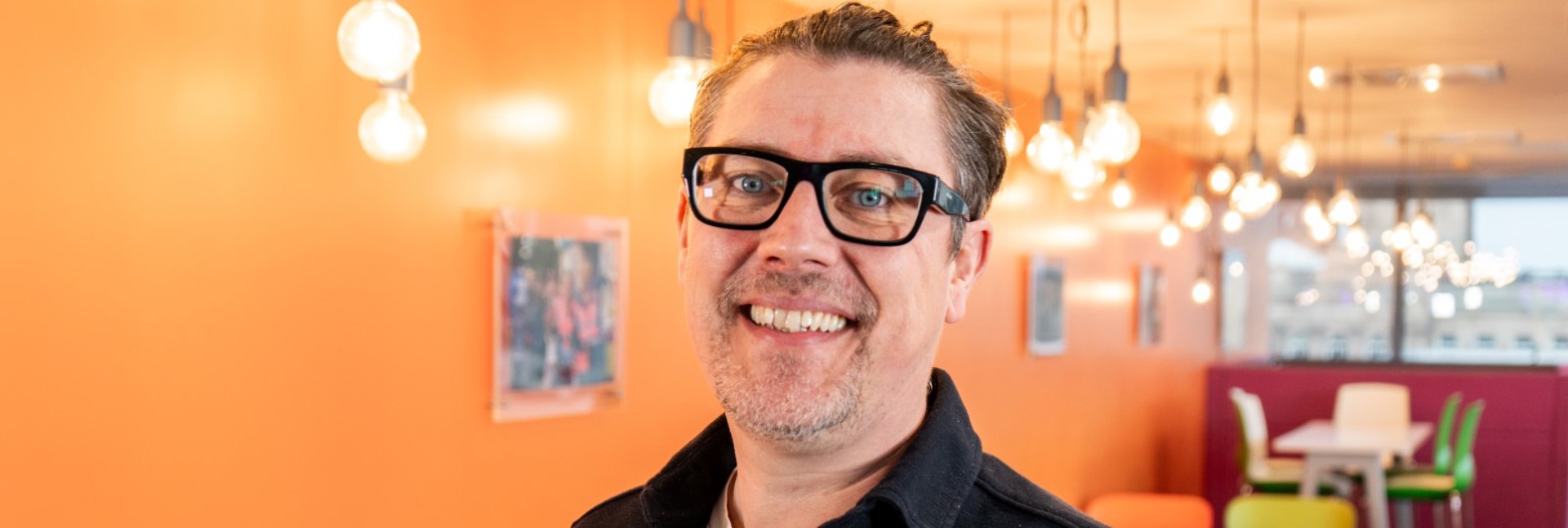Steve Nichol standing in a bright communal area with orange walls and hanging lights.