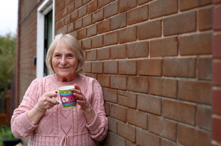 A person in a pink knitted jumper holding a colourful mug while standing beside an outside brick wall.