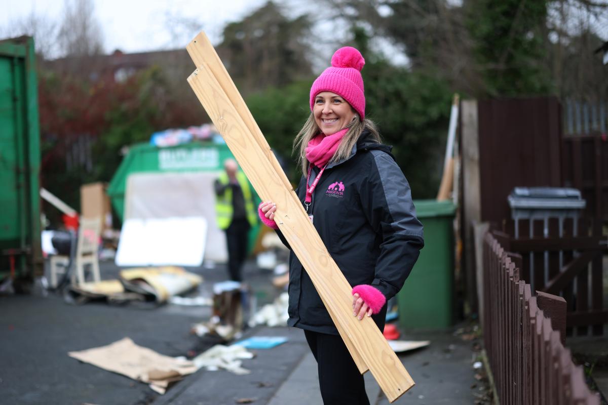A person taking part in a community clean‑up, holding long wooden boards on a street with waste being cleared in the background.